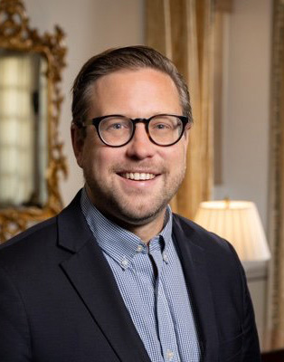 A man wearing glasses and a suit jacket smiles indoors with a lamp and decorative mirror in the background.